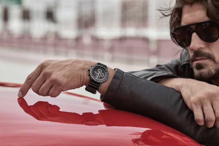 Man leaning on the roof of a car, wearing black JCW aviator sunglasses and the black JCW tachymeter watch.